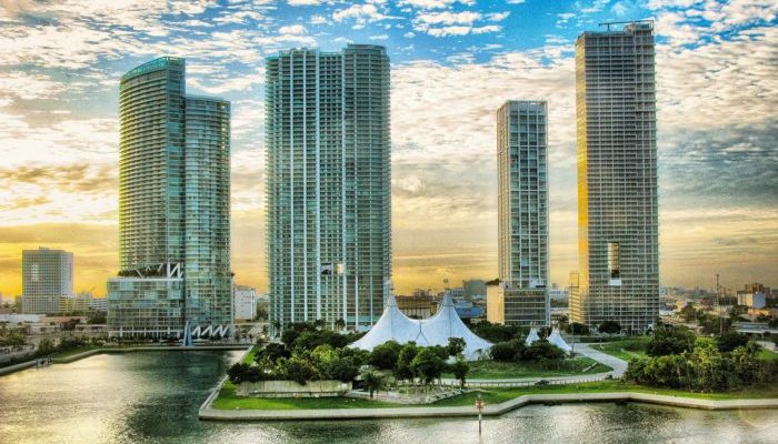 Panoramic view of modern high-rise buildings next to a waterfront with a white pavilion, at sunset, in a cityscape resembling Miami.”